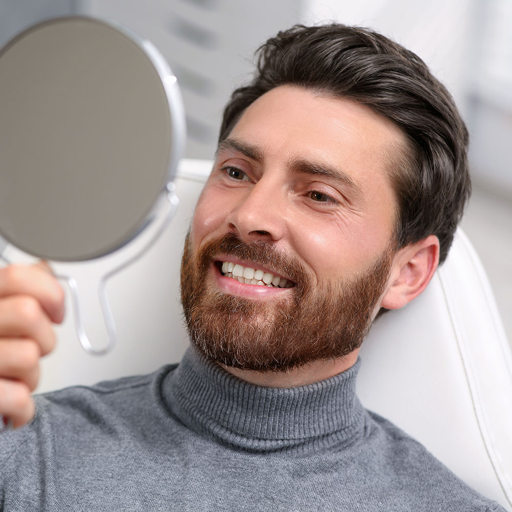 A man with a beard is holding up a mirror to his face, showing both sides of his face.