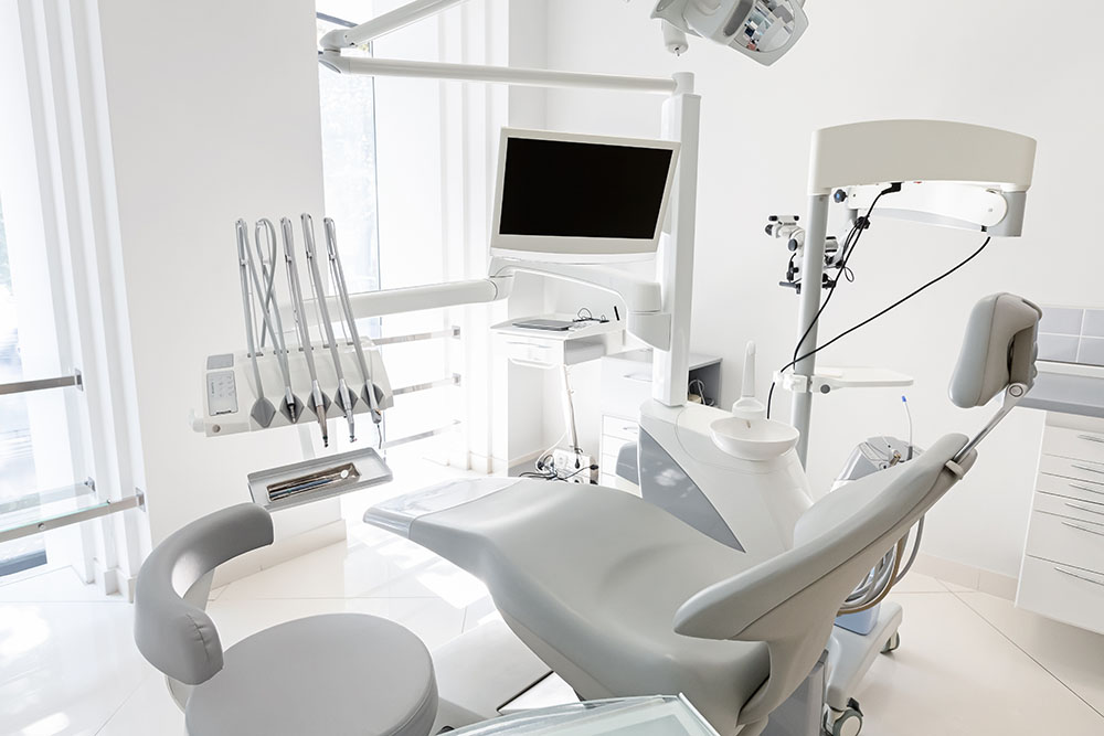 The image shows a modern dental office with a clean and well-equipped interior, featuring a dental chair, examination table, monitor, and various medical equipment.