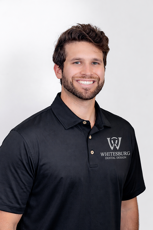 The image shows a man wearing a black polo shirt with a logo on the left side of his chest, a dark-colored lanyard around his neck, and he is smiling at the camera. He appears to be standing in front of a plain background.