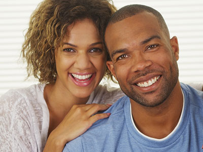 A man and woman posing closely together with smiles, sharing a warm embrace, against a neutral background.