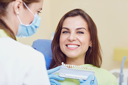The image shows a woman seated at a dental chair, receiving dental care from a professional who is standing behind her, with both smiling and engaged with each other.