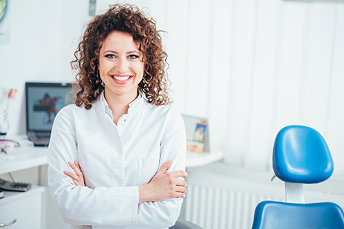 Woman with curly hair, wearing a white shirt, standing in front of dental office chair.
