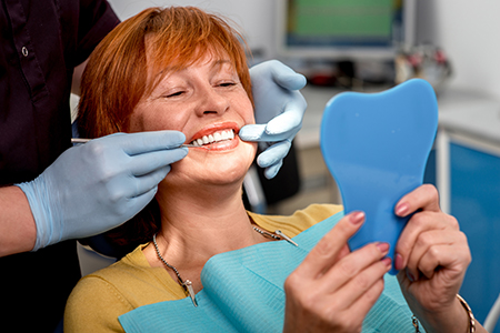 Woman sitting in dental chair with fake teeth held up to her face, smiling at camera while holding toothbrush.