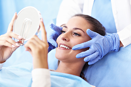 The image depicts a person receiving a medical procedure with a magnifying mirror held over their face by a professional wearing gloves, set against a blue background with a white border at the bottom.