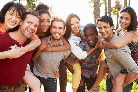 The image features a group of people posing together for a photo, with some individuals hugging each other while smiling at the camera. They appear to be outdoors during daylight hours, under clear skies.