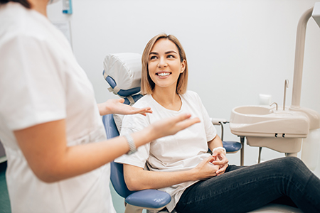 The image shows a woman sitting in a dental chair with her arms crossed, smiling at the camera, while being attended to by a dental professional who appears to be discussing something with her.