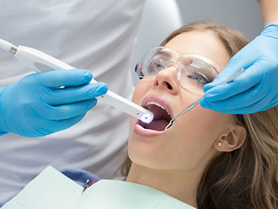 A woman receiving dental care with a dentist using a dental device, wearing gloves and protective eyewear, while other dental staff observe.